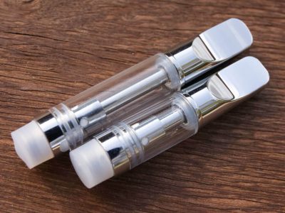 Two Ceramic Cell Vape Cartridges are shown laying lengthwise at an angle against a warm wood textured surface. The cartridges have white mouthpieces and end caps, and a glass body. The longer 1ml cartridge is on top, with the shorter .5ml cartridge below.