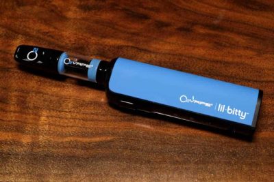 A single blue Lil Bitty Variable Voltage Vape Battery is laid horizontally at an angle against a rich dark wood surface. The mouthpiece is black, with a glass tube and a blue body, with logos on each of the components.