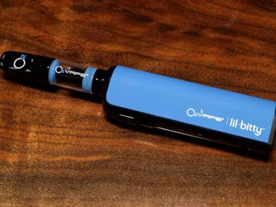 A single blue Lil Bitty Variable Voltage Vape Battery is laid horizontally at an angle against a rich dark wood surface. The mouthpiece is black, with a glass tube and a blue body, with logos on each of the components.