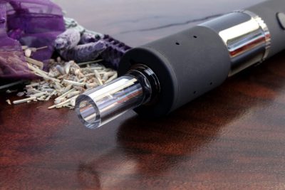 A side angle view of the Dryonic II vape pen with the glass mouthpiece and black body in focus. In the background to the left side is a purple sachet with dry herb spilling out onto the rich dark wood surface.