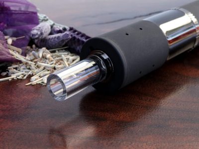 A side angle view of the Dryonic II vape pen with the glass mouthpiece and black body in focus. In the background to the left side is a purple sachet with dry herb spilling out onto the rich dark wood surface.