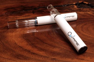 Two All Glass Traveler Extreme vape pens with glass mouthpieces are displayed here, with the 1ml longer cartridge laying horizontally across a warm wood surface, and the shorter .5ml cartridge leaning its glass mouthpiece over the other. The O2VAPE logo is visible at the bottom of the white body.