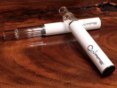 Two All Glass Traveler Extreme vape pens with glass mouthpieces are displayed here, with the 1ml longer cartridge laying horizontally across a warm wood surface, and the shorter .5ml cartridge leaning its glass mouthpiece over the other. The O2VAPE logo is visible at the bottom of the white body.