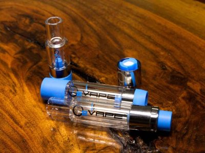2 All Glass Vari-Flow Vape Cartridges are laid horizontally against a warm wood surface. The cartridge is on top is closed, with blue caps at either end. The cartridge beneath and is open. Above the pens, 2 different sized caps are vertically standing at both ends of the cartridges.