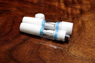2 All Ceramic 510 Vape Cartridges are laid flat against a warm wood surface. The cartridges are white at both ends, with a see-through middle with the O2VAPE logo visible. The cartridge on top is closed. The cartridge beneath is open, with the cap resting vertically above both cartridges.