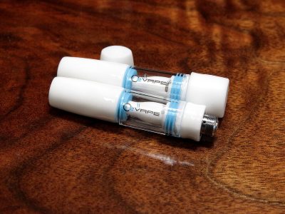 2 All Ceramic 510 Vape Cartridges are laid flat against a warm wood surface. The cartridges are white at both ends, with a see-through middle with the O2VAPE logo visible. The cartridge on top is closed. The cartridge beneath is open, with the cap resting vertically above both cartridges.