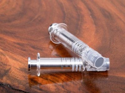 Two glass Dab Syringe Applicators are displayed here. One syringe is laid horizontally across, the other with the end propped up against the horizontal syringe. Gray volumetric indicators are visible along the glass body of both syringes. The metal plunger has a flat handle. The opening of the applicator is visible.