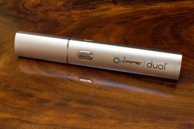 A single steel DUAL Pro Dip & Dab wax vape pen lays horizontally lengthwise against a rich dark wood surface. A power button is located in the center of the body, with the O2VAPE Dual logo on the right side.