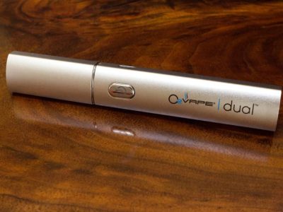 A single steel DUAL Pro Dip & Dab wax vape pen lays horizontally lengthwise against a rich dark wood surface. A power button is located in the center of the body, with the O2VAPE Dual logo on the right side.