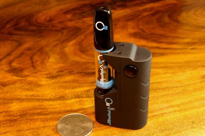 A single Nano Variable Voltage Mini black vape pen stands vertically against a warm wood surface. Laying to the left is a US quarter dollar coin for size comparison. The short side of the pen is 1.5x wider than the coin. The O2VAPE logo is visible on the black mouthpiece, glass tube, and black body. A single button is visible on the body.