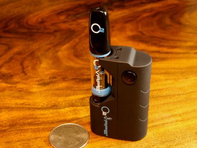 A single Nano Variable Voltage Mini black vape pen stands vertically against a warm wood surface. Laying to the left is a US quarter dollar coin for size comparison. The short side of the pen is 1.5x wider than the coin. The O2VAPE logo is visible on the black mouthpiece, glass tube, and black body. A single button is visible on the body.