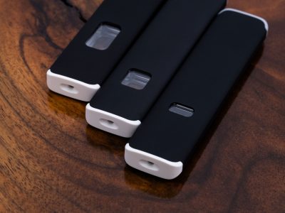 3 black Aero Plus Slim Vape Pens of varying capacities are arranged side by side and slightly offset with the short white mouthpiece ends in focus. A translucent window into the pen's contents is visible on the flat side of the pens.