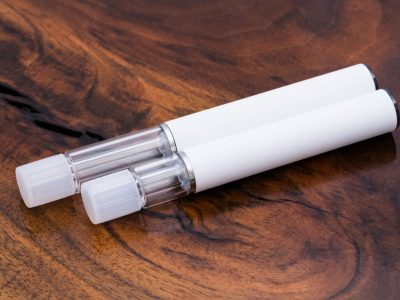Two Traveler Extreme® All Glass Disposable Vape Pens are resting diagonally on a rich wood surface. The pens are sleek and cylindrical, with a white body over its all-glass construction. The pen in the front is shorter and has a .5ml capacity, while the one in the background is longer and has a 1ml capacity. There are caps inserted into the mouthpieces.