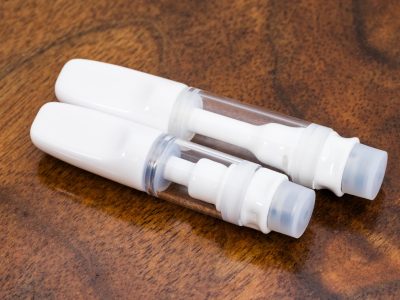 Two white Full Ceramic vape cartridges with glass bodies are laid horizontally at an angle over a warm wood surface. The taller 1ml cartridge is on top, with the shorter .5ml cartridge beneath.