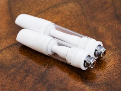 Two All Ceramic 510 Vape Cartridges are laid at an angle side by side against a warm wood surface. The mouthpiece ends are on the left, with the screw side on the right. The longer 1ml cartridge is on top while the shorter .5ml cartridge is beneath.