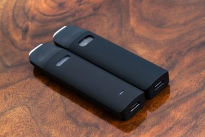 Two black Traveler Extreme ® vape pens are laid flat at an angle against a warm wood surface. A small translucent cartridge window is located on the flat side of the body. At far end is the protruding mouthpiece, with the USB-C charging port at the bottom end.