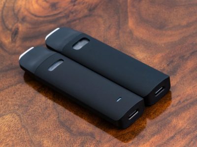 Two black Traveler Extreme ® vape pens are laid flat at an angle against a warm wood surface. A small translucent cartridge window is located on the flat side of the body. At far end is the protruding mouthpiece, with the USB-C charging port at the bottom end.